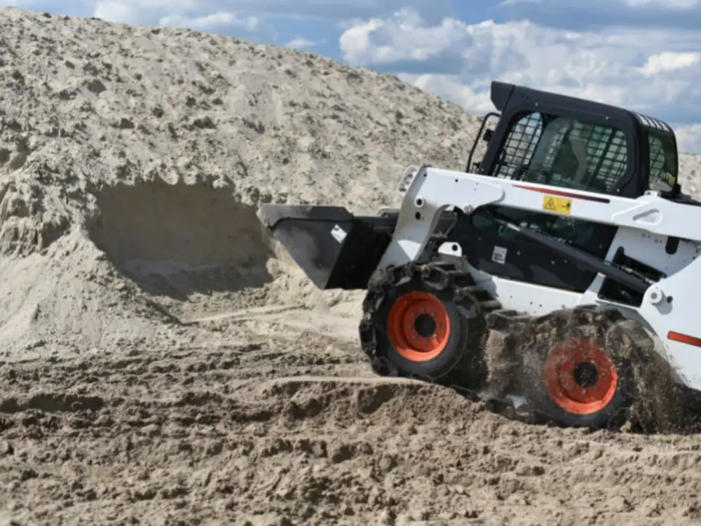 Mini cargador compacto Bobcat blanco con ruedas naranjas, moviendo una gran pila de arena en una obra. Maquinaria de Escombros Áridos trabajando con eficiencia en movimiento de tierras.