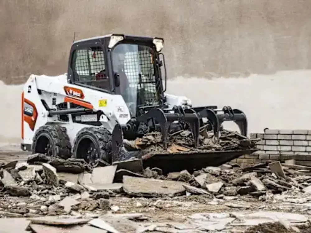 Mini cargador compacto Bobcat blanco y naranja con un cucharón de garra para demolición, recogiendo y manejando escombros de construcción en una zona urbana o de obra. Maquinaria de Escombros Áridos para gestión y remoción de desechos de demolición.