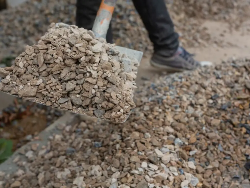 Primer plano de una pala metálica llena de áridos de construcción de Escombros Áridos. Se aprecian las texturas y colores de la grava, con un trabajador de fondo en la zona de acopio.