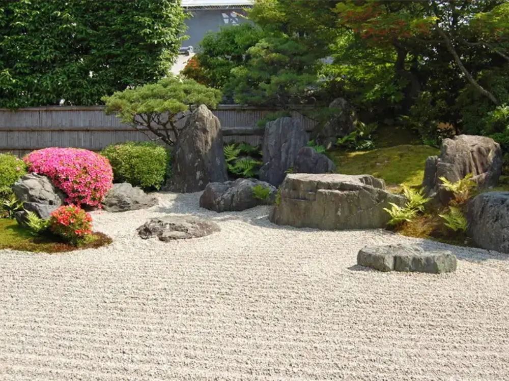 Paisaje de un jardín zen japonés de Escombros Áridos, mostrando gravilla fina de color claro rastrillada para simular agua, con rocas grandes que representan islas, musgo y un arbusto de azaleas rosadas.