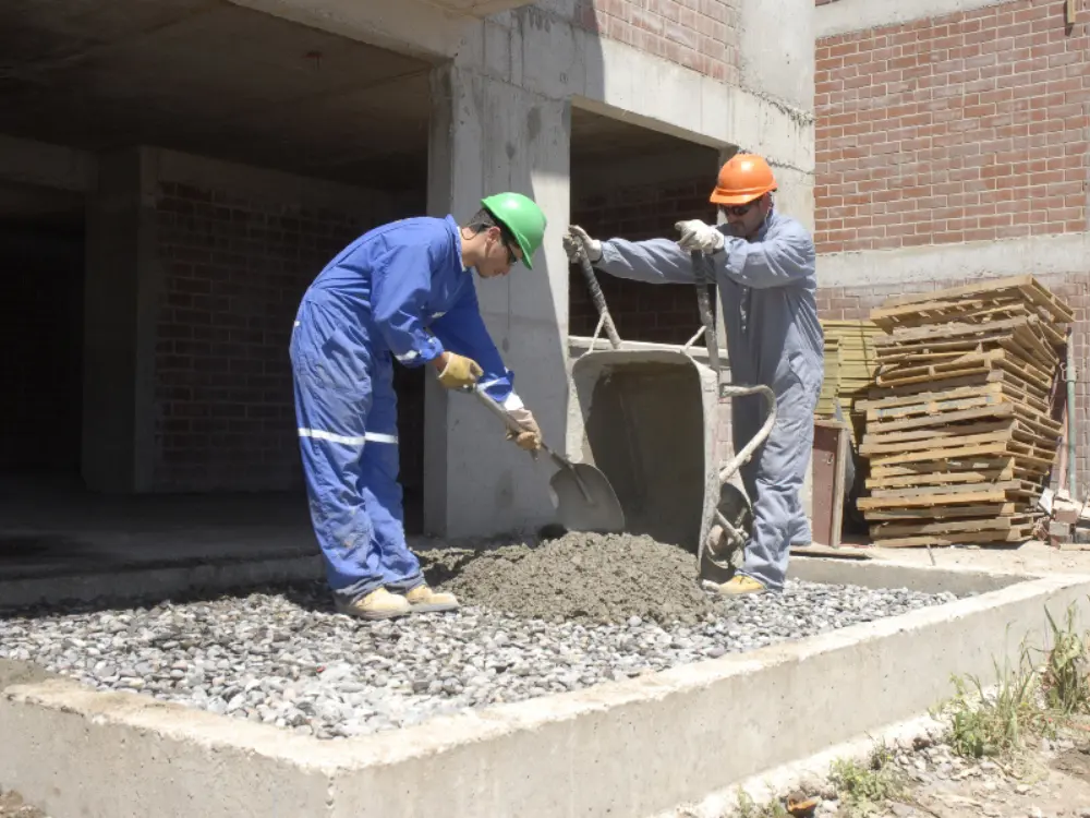 Dos trabajadores de la construcción preparando hormigón a mano. Uno de ellos usa una pala sobre una mezcla que contiene gravilla 3/4, demostrando el uso de áridos de calidad para cimientos y estructuras, como los que vende y despacha Escombros Áridos.