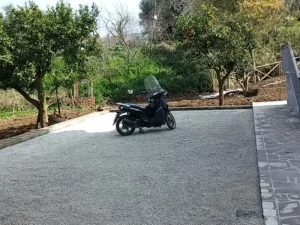 Vista de un área de estacionamiento o acceso en un entorno ajardinado, cubierta con grava estabilizada de color claro (gris/beige). Una scooter está aparcada en la superficie, mostrando la firmeza del terreno. El árido utilizado es suministrado por Escombro Áridos, garantizando un acabado limpio, transitable y permeable.