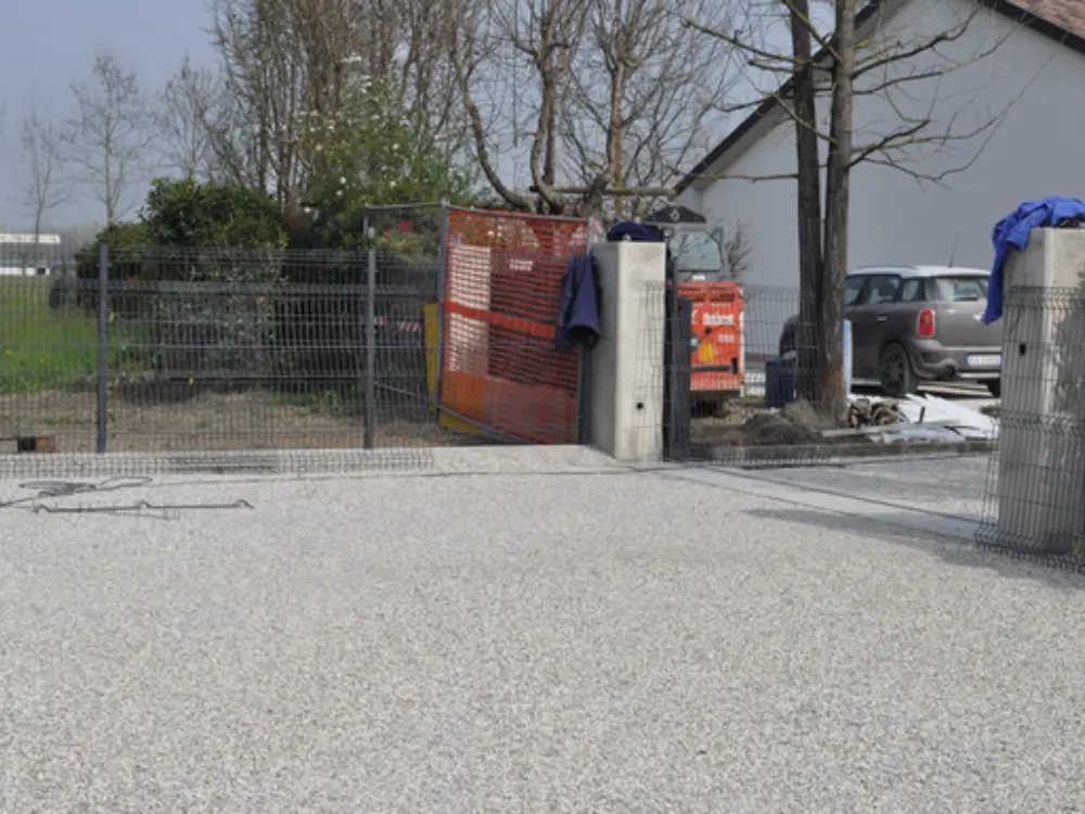 Vista de una entrada de propiedad residencial en fase de construcción, con la superficie ya finalizada usando grava estabilizada de color blanco o gris muy claro. Se aprecian los pilares de un futuro portón y una rejilla de cercado, demostrando la aplicación del árido de Escombro Áridos como una base firme y permeable ideal para accesos vehiculares.