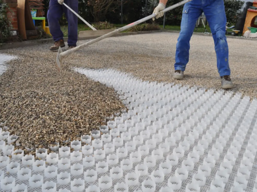 Primer plano del proceso de extendido y nivelación de grava decorativa (árido marrón/beige) sobre una malla estabilizadora de plástico blanca. Se ven dos trabajadores utilizando rastrillos para rellenar las celdas de la rejilla. Este sistema asegura un pavimento firme y drenante utilizando áridos de calidad suministrados por Escombro Áridos.