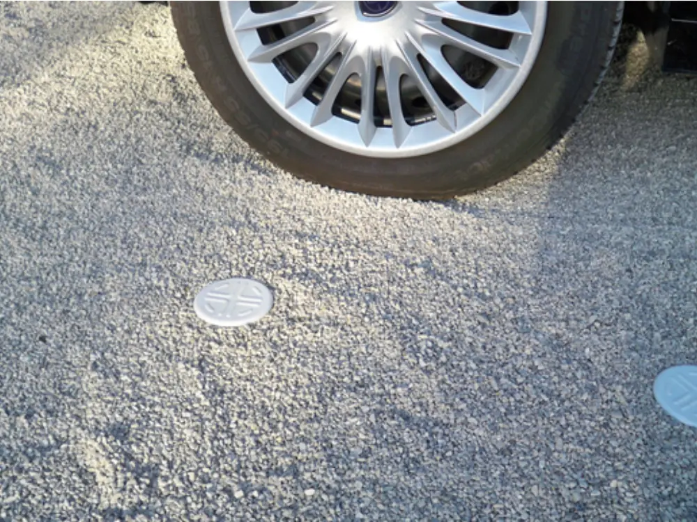 Primer plano que muestra el detalle de una superficie de grava estabilizada de color gris claro, con la rueda y el rin de un automóvil parcialment visible. Se aprecian pequeños tapones circulares de color claro, probablemente parte del sistema de rejilla estabilizadora, que aseguran la firmeza del terreno bajo el peso del vehículo. El árido utilizado es de alta resistencia y es suministrado por Escombro Áridos.