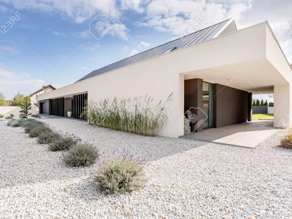 Fachada de villa moderna con diseño minimalista que utiliza gravilla blanca decorativa en el jardín. El material se extiende a lo largo de la entrada y se complementa con plantas de lavanda y pastos. Escombros Áridos vende esta gravilla para paisajismo moderno.
