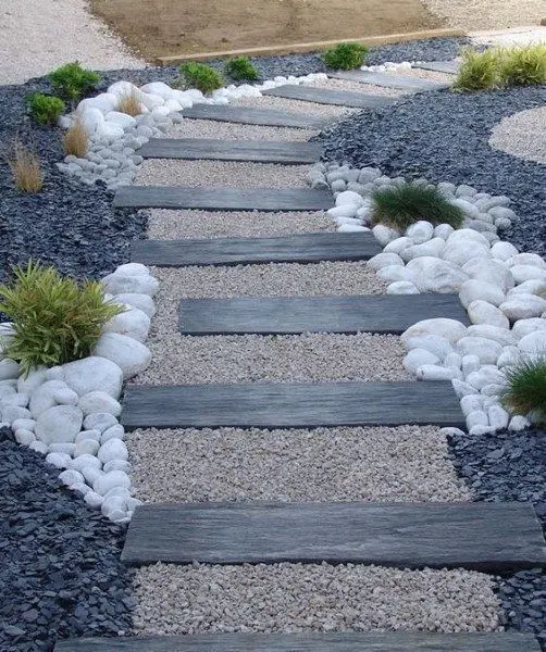 Diseño de sendero moderno con escalones de piedra o madera, rodeado por gravilla fina beige en el centro, guijarros blancos decorativos y gravilla oscura en los bordes. Escombros Áridos ofrece venta de gravilla para diseños de paisajismo en Santiago.
