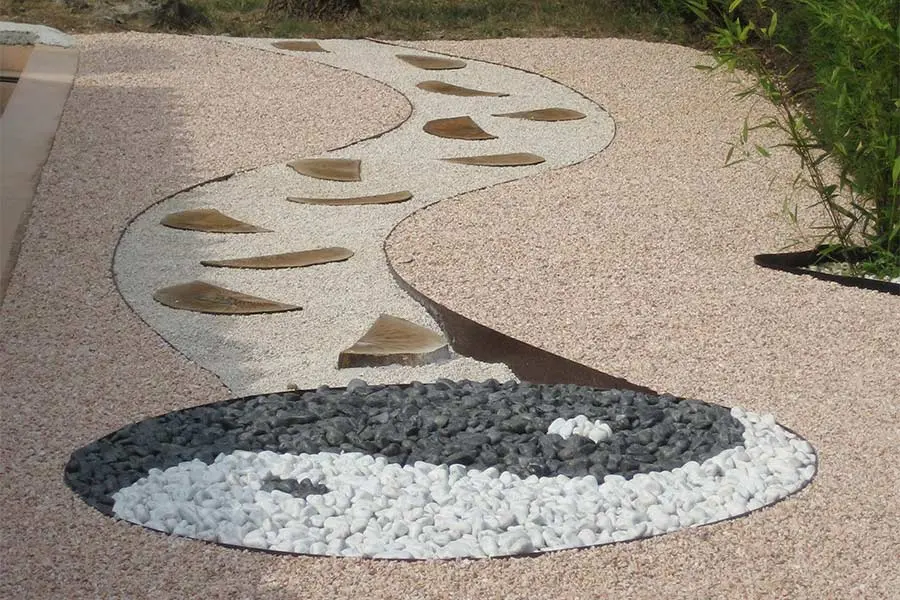 Jardín decorativo con sendero curvo de gravilla blanca y rosa, complementado con un diseño Yin Yang de piedras blancas y grises, destacando la versatilidad de los productos de Escombros Áridos.