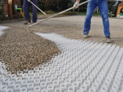 Primer plano del proceso de extendido y nivelación de grava decorativa (árido marrón/beige) sobre una malla estabilizadora de plástico blanca. Se ven dos trabajadores utilizando rastrillos para rellenar las celdas de la rejilla. Este sistema asegura un pavimento firme y drenante utilizando áridos de calidad suministrados por Escombro Áridos.