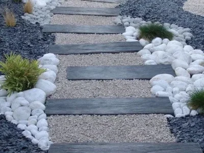 Diseño de sendero moderno con escalones de piedra o madera, rodeado por gravilla fina beige en el centro, guijarros blancos decorativos y gravilla oscura en los bordes. Escombros Áridos ofrece venta de gravilla para diseños de paisajismo en Santiago.