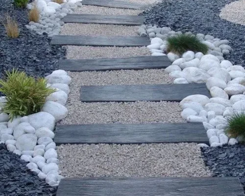 Diseño de sendero moderno con escalones de piedra o madera, rodeado por gravilla fina beige en el centro, guijarros blancos decorativos y gravilla oscura en los bordes. Escombros Áridos ofrece venta de gravilla para diseños de paisajismo en Santiago.