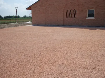 Superficie de grava estabilizada de color terracota (rojizo claro) instalada como zona de aparcamiento o acceso frente a una construcción de ladrillo. Esta solución utiliza áridos de alta resistencia de Escombro Áridos para crear un acabado drenante, firme y estético.